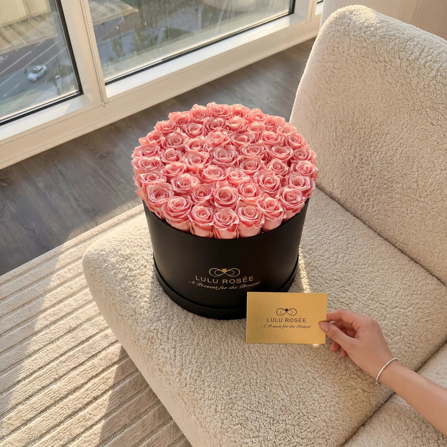 LULU ROSÉE extra large round box baby pink roses black classic box on a beige couch near a window.