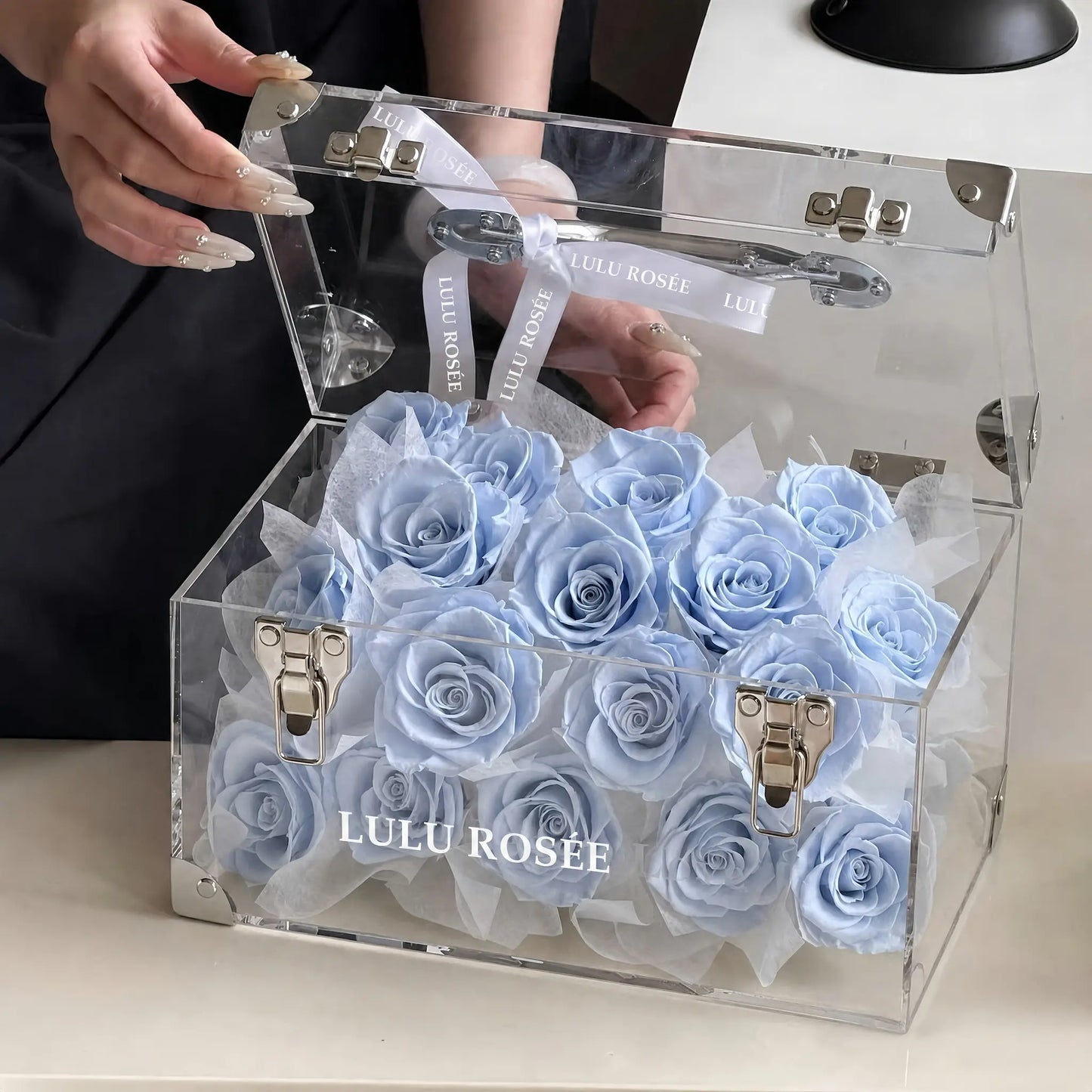 LULU ROSÉE Petite Eternal Acrylic Handbag Rose Box, small transparent acrylic handbag-shaped gift box filled with soft baby blue preserved roses with open lid, handheld lifestyle shot, customizable flower color options, mini luxury everlasting floral souvenir for birthday, anniversary & romantic gift giving